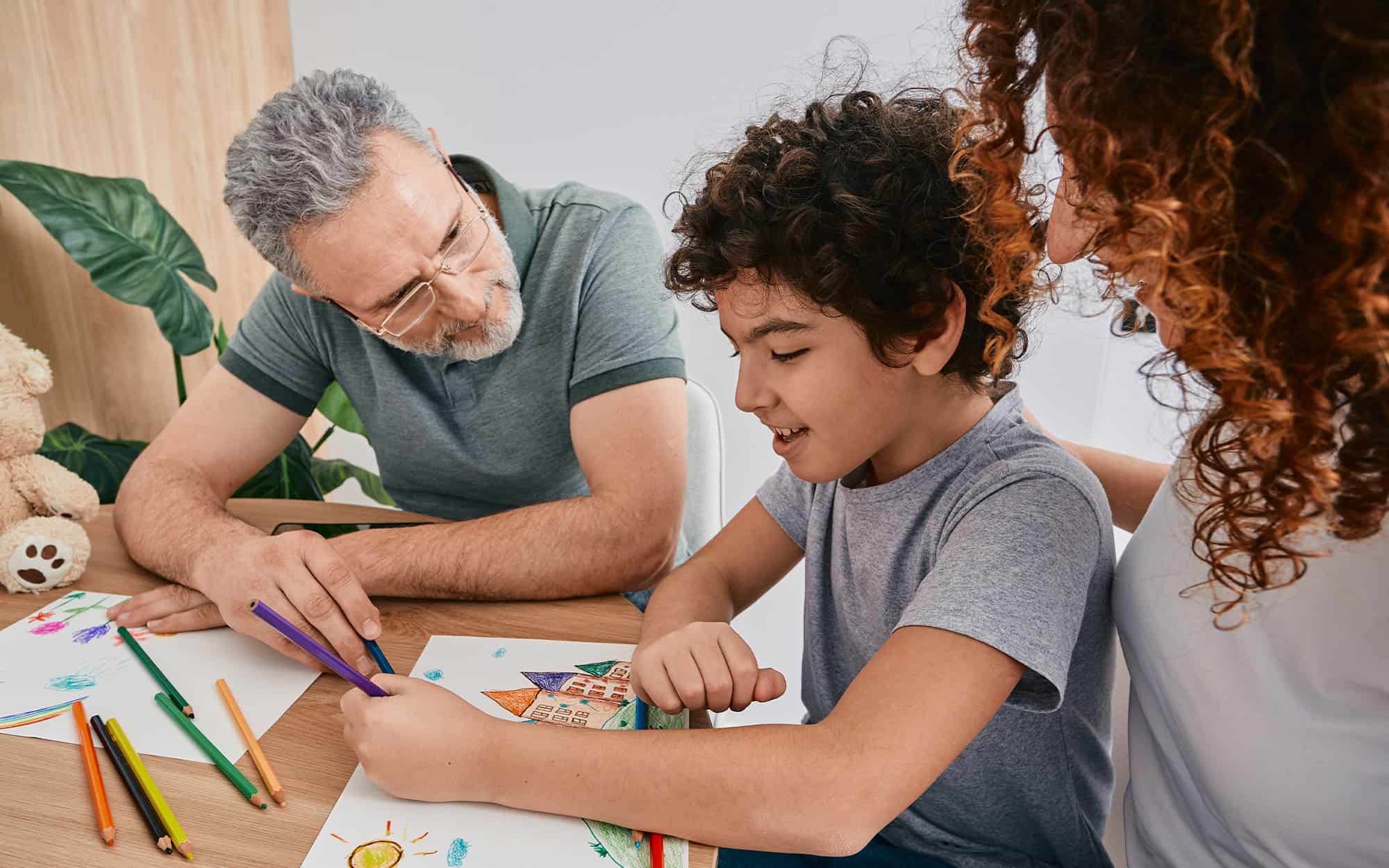 Side view of a child coloring while two adults are standing nearby