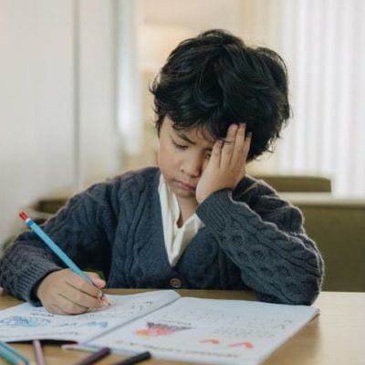 A little Asian boy resting his head on his arms as he sits at his desk looking bored