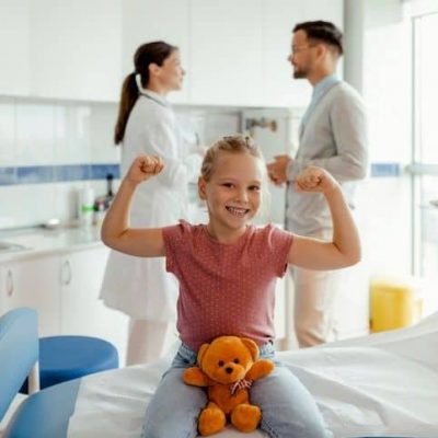 Happy Little Girl With Teddy Bear Flexing Her Muscles After Medical Examination at Doctor's Office While Doctor Talking to Her Father on Background