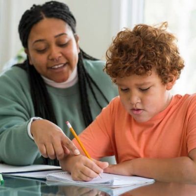 Beautiful mother is smiling gently as she is helping her elementary age son with his homework from school at the kitchen table. She is pointing at a mistake on the page and he is using a pencil to correct the work in the notebook.
